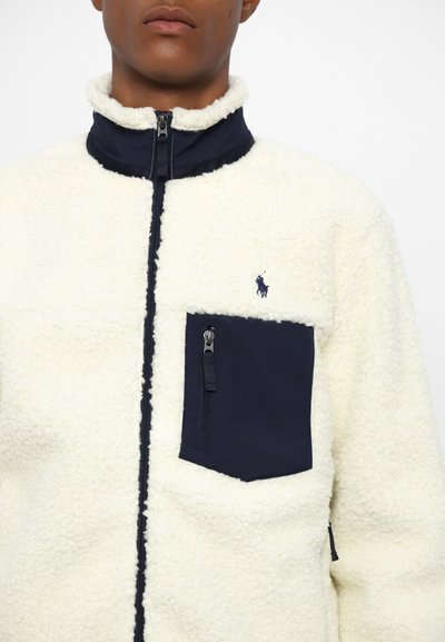 Fleece jacket featuring a cream body, navy collar, and chest pocket. Zip closure, soft texture, and a small embroidered logo on the chest.