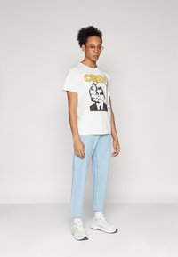 Λευκό γραφικό μπλουζάκι με κείμενο "OBEY" και σχέδιο χεριού. Ανοιχτό μπλε τζιν και λευκά αθλητικά παπούτσια. Αξεσουάρ περιλαμβάνουν ροζ γυαλιά ηλίου.