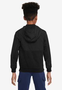 Schwarze Kapuzenjacke mit einem Reißverschluss, die ein schlichtes Design und elastische Bündchen aufweist, von hinten an einer Person mit blauen Hosen gesehen.