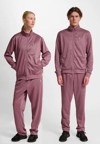 Trainingspakken van zachte mauvekleurige stof. Beide hebben hoge kragen, ritsen aan de voorkant, elastische manchetten en bijpassende broeken met zakken. Zwarte sneakers gedragen.