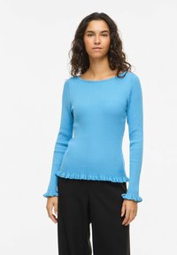 Femme portant un pull bleu ajusté à manches longues, côtelé, avec des poignets et un ourlet volantés, associé à un pantalon noir ample, debout devant un fond blanc.