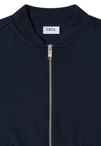 Veste bleu marine avec fermeture éclair avant argentée et petite étiquette blanche portant l'inscription "CECIL" au col du cou.