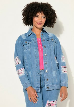 Veste en denim bleu clair avec des déchirures sur les manches, des boutons en argent et deux poches poitrine. Portée par-dessus un haut rose vif.