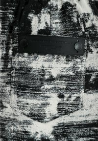 Tela texturizada en blanco y negro que presenta un bolsillo con una etiqueta de cuero negra y broches redondos. El patrón parece abstracto con sombras variadas.