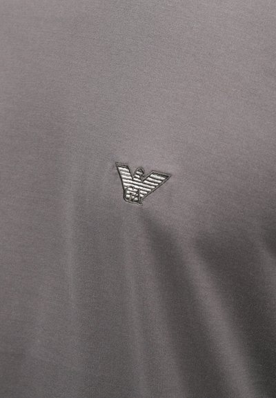 Pelēks T-krekls, kas izgatavots no gludas auduma, ar nelielu izšūtu ērgļa logotipu augšējā kreisajā pusē. Vienkāršs un minimālistisks dizains.