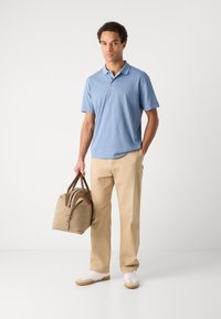 Polo shirt albastru deschis, pantaloni bej și adidași albi. Ținând o geantă de voiaj bej cu accente maro și fermoar. Design simplu.