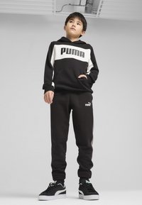 Hoodie PUMA noir et blanc avec poche frontale, pantalon de jogging noir et baskets noires avec des accents blancs et verts, le tout sur fond uni.