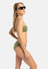 Olive žalios spalvos bandeau tipo bikini viršus ir atitinkamos kelnaitės, pagamintos iš lygaus audinio. Modelis dėvi juodus akinius nuo saulės ir turi banguotus plaukus.