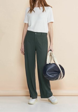 Vrouw met een wit topje met korte mouwen, donkergroene wijde broekspijpen, witte sneakers, en een zwarte en beige ronde duffeltas in haar hand.