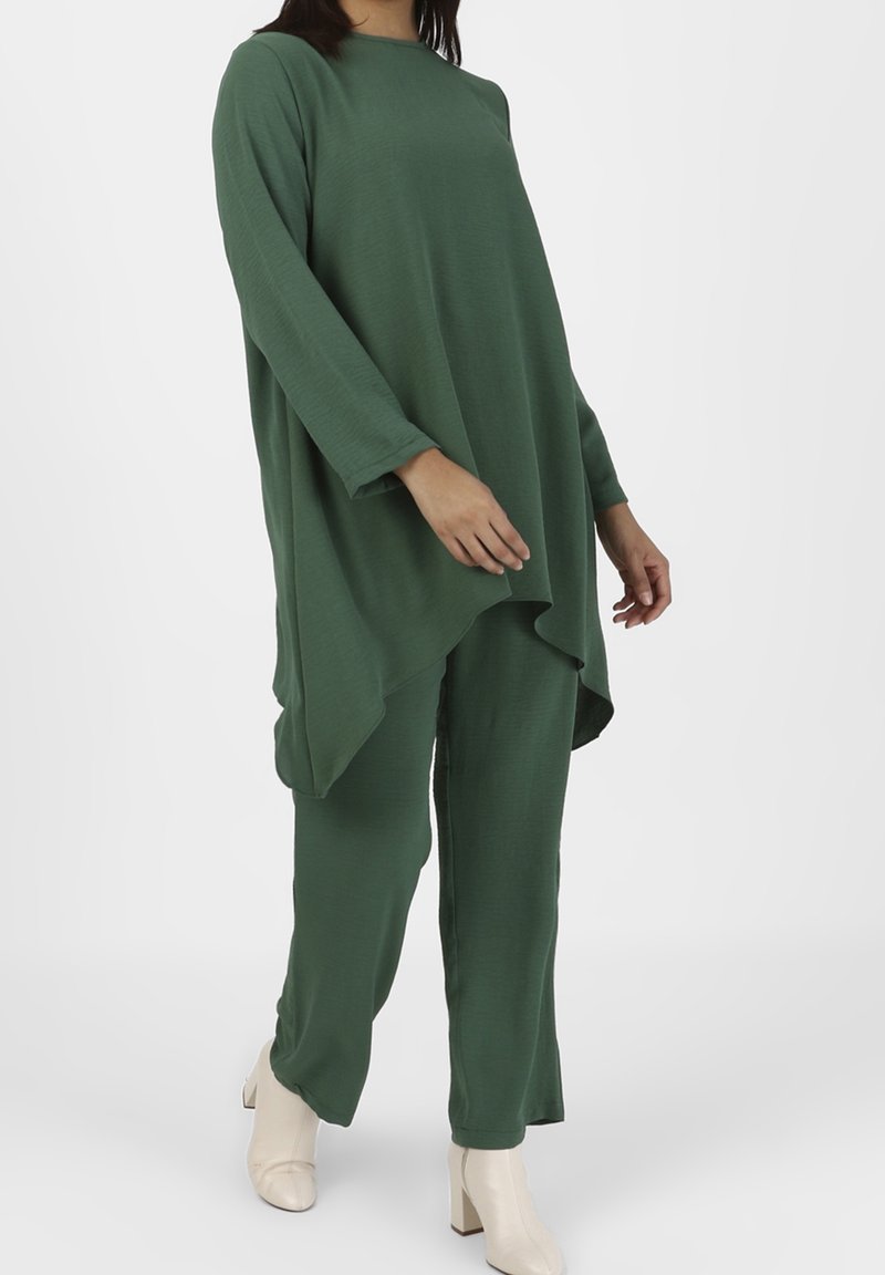 Femme portant une tunique ample à manches longues vert foncé avec un pantalon large assorti et des bottines blanches à talons.