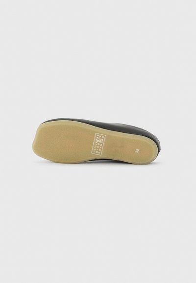 Μαύρο παπούτσι με γκριζωπό καφέ επίπεδη σόλα στραμμένη προς τα επάνω, μέγεθος 35 σημειωμένο στη φτέρνα και λευκή ετικέτα πληροφοριών προϊόντος κοντά στην καμάρα.