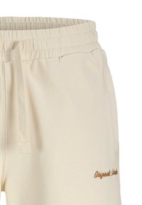 Pantalon de survêtement beige avec taille élastique et cordon, arborant un texte brodé marron sur la partie inférieure de la jambe droite. Texture de tissu lisse.