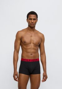 Μαύροι μπόξερ με κόκκινη μέση που φέρει την επιγραφή "HILFIGER". Μαλακό ύφασμα, εφαρμοστή σχεδίαση, μήκος μέχρι το μέσο του μηρού. Το μοντέλο στέκεται με αυτοπεποίθηση.