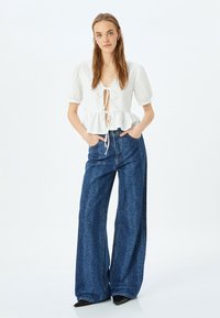 Top bianco corto con maniche a palloncino e dettaglio con nodo frontale, abbinato a jeans blu a vita alta e gamba larga con un motivo sottile.