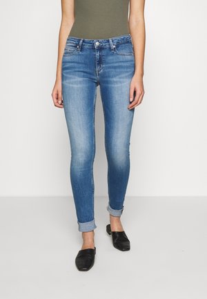 Kobieta ubrana w jasnoniebieskie obcisłe jeansy z podwiniętymi nogawkami, oliwkowozielony bezrękawnik i czarne teksturowane buty wsuwane, stojąca na jasnej podłodze.