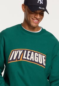Felpa verde con una grande grafica "IVY LEAGUE" in crema e arancione. Presenta un collo a giro e una texture morbida. Indossata con un cappellino nero.