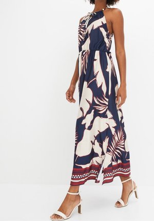 Femme portant une robe maxi sans manches bleu marine à fleurs blanches avec une bordure rouge et des sandales blanches à talons, debout devant un fond uni.