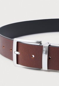 Καφέ δερμάτινη ζώνη με λεία υφή και ασημένια μεταλλική αγκράφα. Διαθέτει ρυθμιζόμενες τρύπες και λογότυπο κοντά στην αγκράφα.