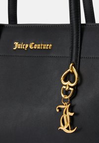 Melnas mākslīgās ādas rokassomiņa ar zelta "Juicy Couture" logotipu, sirds formas aksesuāriem un dekoratīvu zelta piekariņu. Gluda tekstūra.