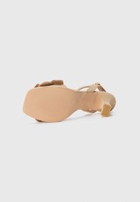 Beige hoge hakken sandaal met een gevoerde zool, open achterkant en slanke banden. Inclusief een gestructureerde buitenzool en een kleine blokhak.