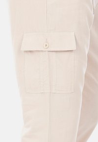 Pantaloni cargo di un beige chiaro con una grande tasca a soffietto sulla coscia, caratterizzati da una chiusura a bottone e dettagli cuciti. Tessuto dalla finitura liscia.