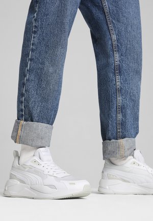 Zapatillas atléticas blancas con un diseño texturizado, que cuentan con una suela gruesa, parte superior de tela de malla y detalles reflectantes. Usadas con jeans azules de mezclilla con los bajos doblados.