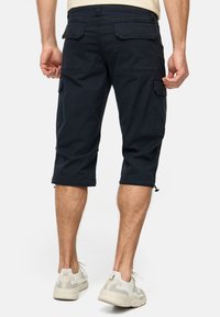 Shorts cargo navy en mélange de coton, comprenant six poches, de coupe à hauteur du genou, et avec des hémis à cordon. Portés avec des baskets de couleur claire.