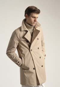 Gabardina beige de doble botonadura con grandes botones marrones, cuello con muesca y bolsillos laterales, combinada con un suéter de punto.