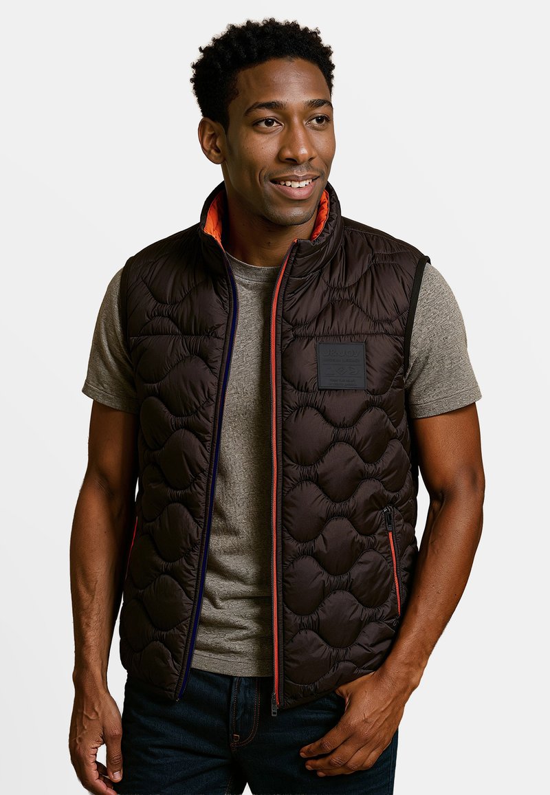 Dikke zwarte vest met oranje binnenkant, voorzien van een ritssluiting, zijzakken en een gestructureerd gewatteerd patroon. Draagt over een grijs T-shirt.