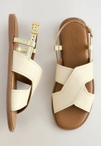 Cremefarbene Leder-Sandalen mit Überkreuz-Design, goldenen Riemen und braunem gepolstertem Fußbett, ausgestattet mit einer Schnallenverschluss an dem Knöchelriemen.
