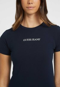 Camiseta de algodón azul marino con mangas cortas, que presenta el logo "GUESS JEANS" en texto blanco en el pecho. Textura suave, diseño ajustado.