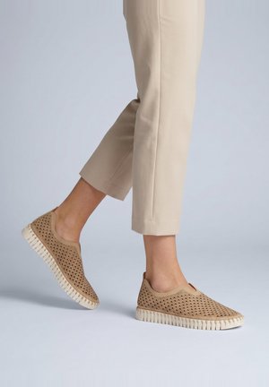 Persona que lleva zapatos beige sin cordones con partes superiores estampadas y suelas blancas, combinados con pantalones beige claro recortados sobre un fondo liso.