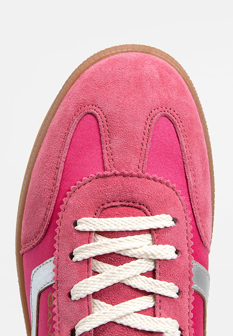 Zapatilla rosa con una combinación de materiales de ante y tela, cordones blancos y un acento plateado. La suela es marrón claro y texturizada.