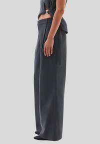 Person som bär mörkgrå vida byxor och en kort topp med ett sidofäste, stående på sidan mot en enfärgad bakgrund.