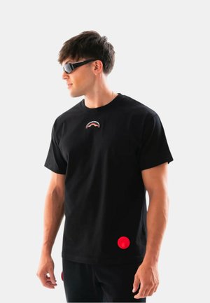 Giovane uomo con maglietta nera con piccolo logo di bocca di squalo sul petto e occhiali da sole neri, in piedi davanti a uno sfondo bianco semplice.