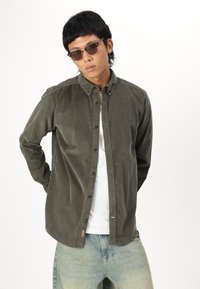 Chemise en velours côtelé vert avec un design à boutons, manches longues et un ourlet légèrement arrondi, portée par-dessus une chemise blanche. Les lunettes sont foncées et arrondies.