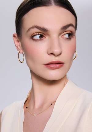 Złote kolczyki w kształcie kół oraz delikatny naszyjnik z czarnymi i białymi koralikami, noszone z kremowym, teksturowanym ubraniem.