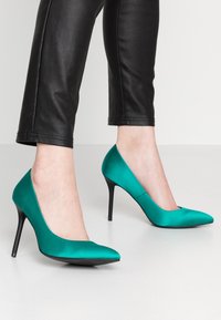 Tyrkysové saténové lodičky s špičatou špičkou a tenkými černými stiletto podpatky, spárované s černými upnutými koženými kalhotami. Hladká textura.