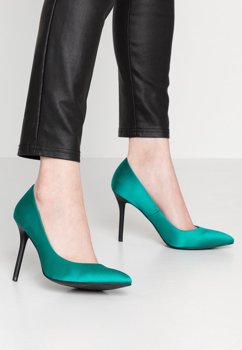 Tyrkysové saténové lodičky s špičatou špičkou a tenkými černými stiletto podpatky, spárované s černými upnutými koženými kalhotami. Hladká textura.