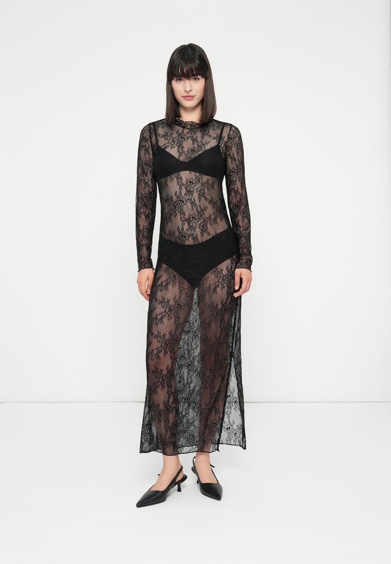 Žena oblečená v průhledných černých krajkových šatech s dlouhým rukávem, pod kterými má černou podprsenku a kalhotky s vysokým pasem, stojí před bílým pozadím.