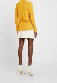 Gelber Strickpullover mit hohem Kragen, gerippten Bündchen und Saum. Getragen über einem kurzen cremefarbenen Rock und kombiniert mit cremefarbenen Stiefeletten.