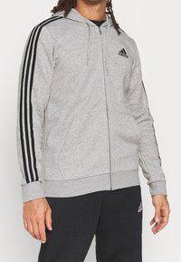 Sudadera gris con cremallera y capucha, con estampados en negro en los hombros y bolsillos frontales. Fabricada en un tejido suave y texturizado. Logo de adidas en el pecho.