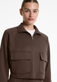 Vrouw in een bruine zip-up jas met grote klepzakken over een witte blouse, met het gezicht naar voren en een neutrale uitdrukking.