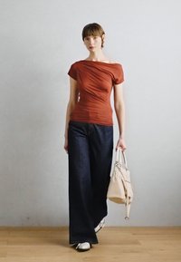 Haut ajusté de couleur rouille avec un design dénudé d'épaule, associé à un jean en denim foncé à jambe large et un sac à main crème. Texture simple et lisse.