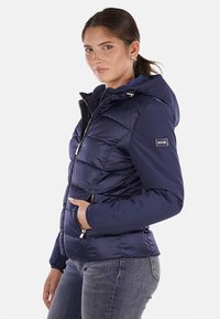 Giacca imbottita blu navy con zip frontale, cappuccio e tasche laterali. Presenta un design trapuntato e una patch con logo sulla manica. Tessuto morbido.
