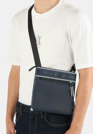 Uomo che indossa una T-shirt bianca Armani Exchange e jeans scuri, con una borsa a tracolla nera con cerniera e tracolla con marchio.