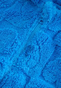 Veste douce et bleu vif avec une surface texturée et pelucheuse, dotée d'une fermeture éclair à l'avant. Présente un motif doux et nuageux sur l'ensemble du tissu.