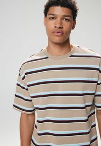 Kortærmet, beige stribet t-shirt med vandrette striber i lys blå, sort og mørkerød. Klassisk crew neck-design.