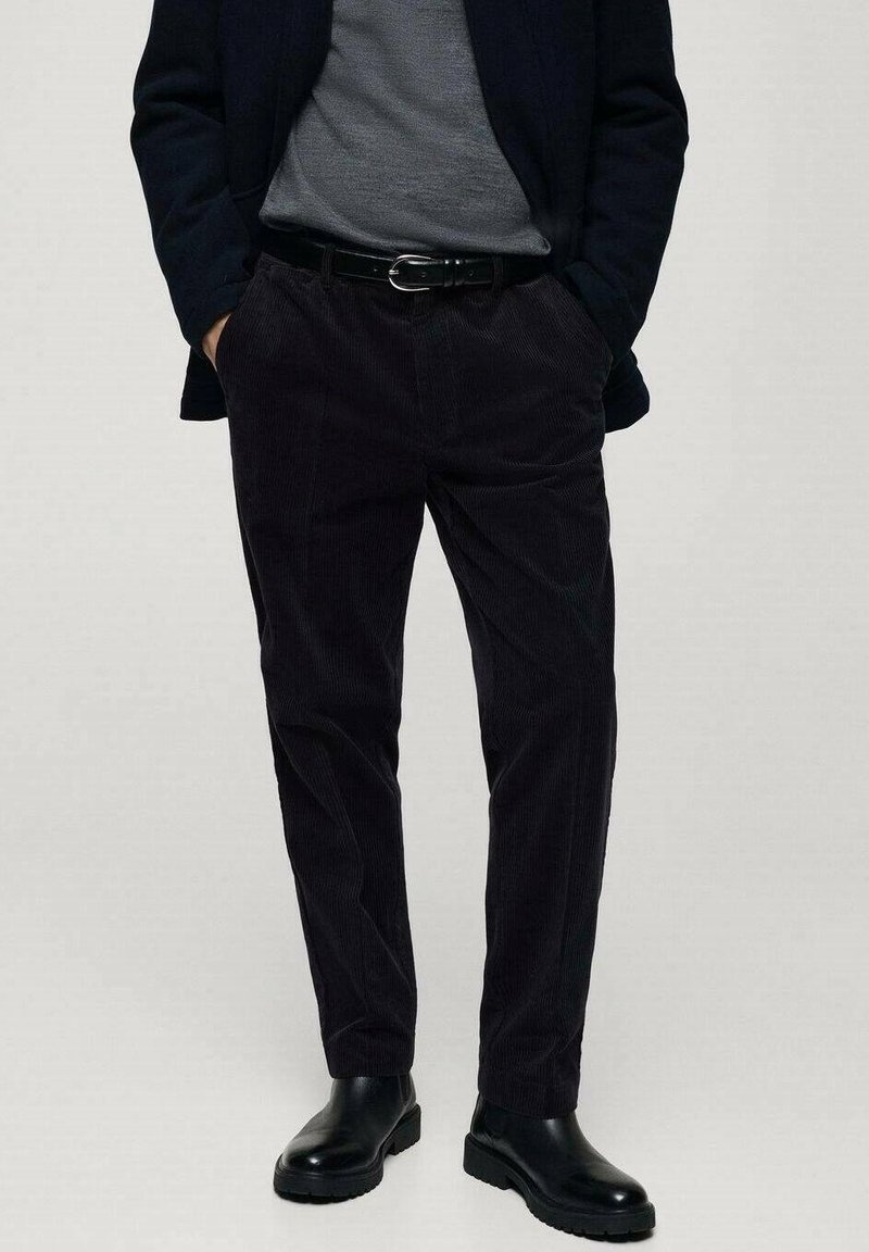 Homme portant un pantalon en velours côtelé noir, une ceinture en cuir noire, des bottes noires, un pull gris et une veste noire les mains dans les poches.