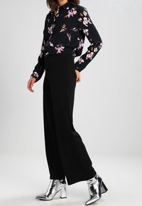 Pantaloni neri a gamba larga abbinati a una camicetta a maniche lunghe con fantasia floreale. Il modello indossa stivaletti metallici argentei alla caviglia.